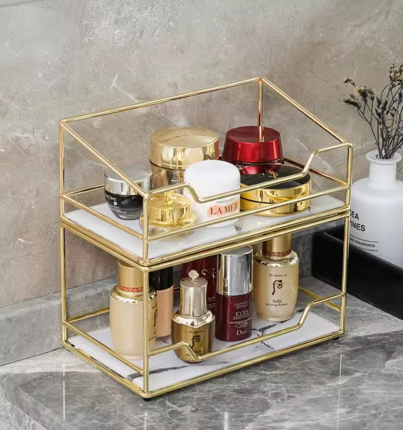 Gold metal desktop makeup organizer rack with white shelves holding various cosmetic bottles