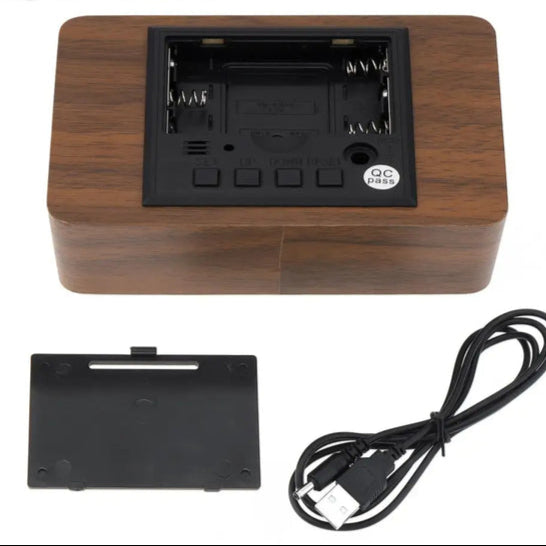 Back view of a digital wooden alarm clock showing battery compartment cover and USB charging cable