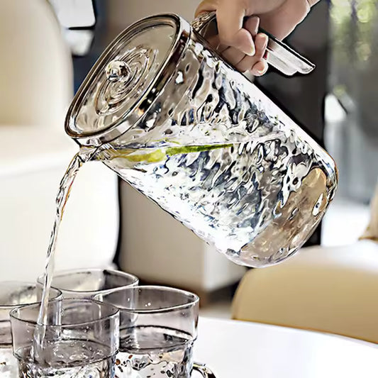 Clear glass juice pitcher with lid being used to pour water into glasses on a table