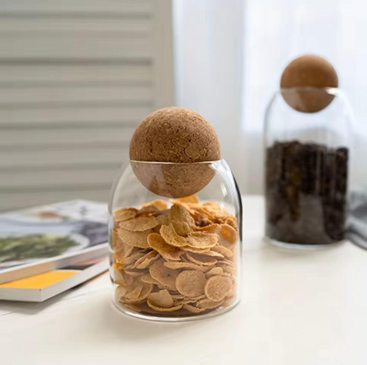 Clear glass storage jars with cork lids holding cereal flakes and coffee beans on a white table
