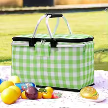 Green and white checkered large foldable picnic basket on outdoor blanket with fruits and snacks