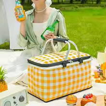 Large foldable picnic basket in yellow checkered pattern with handles on grass next to a person holding drinks