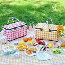 Large foldable picnic basket in red and yellow checkered patterns with handles on a picnic blanket with food and drinks