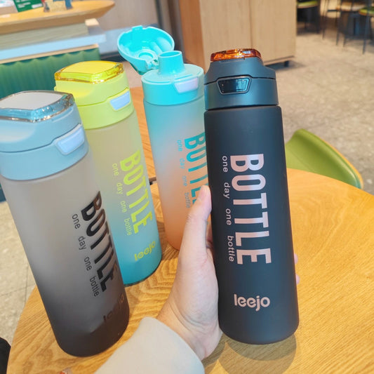 Hand holding black 1000ml sports water bottle with other colorful bottles on wooden table