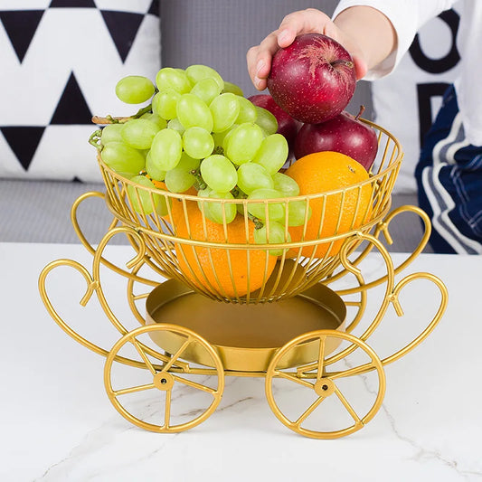 Gold carriage style modern fruit basket filled with green grapes, oranges, and red apples on white table