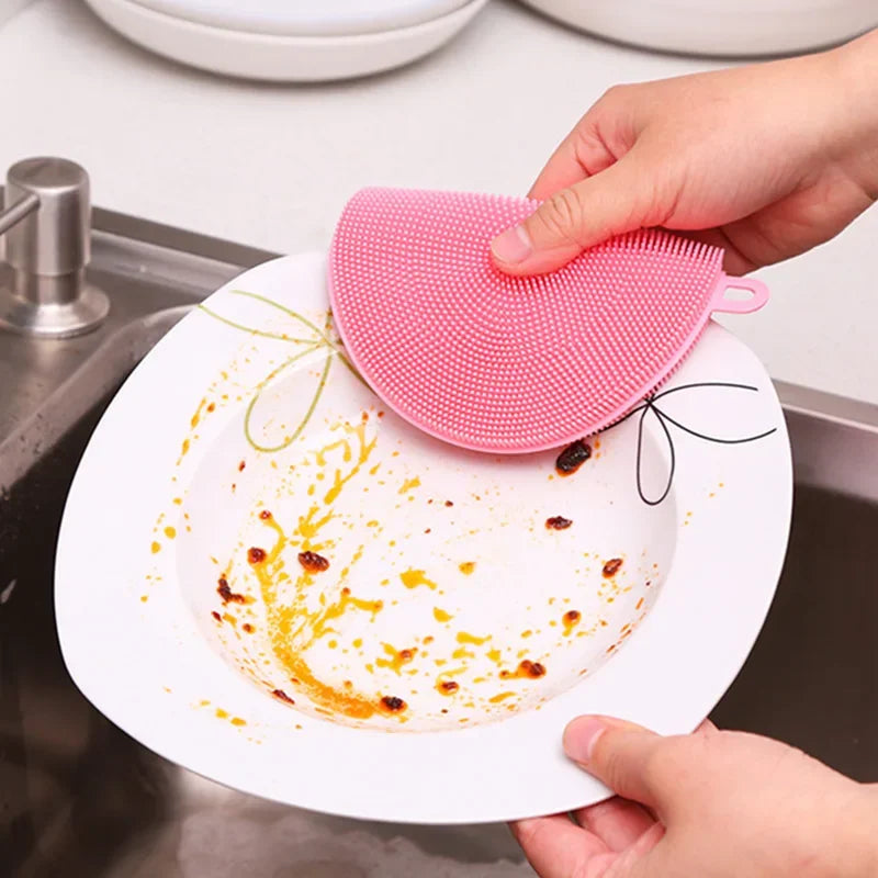 Pink multi-functional kitchen scrubber cleaning a dirty white plate over a kitchen sink