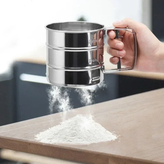 Hand holding stainless steel flour sieve sifting flour onto wooden countertop in kitchen