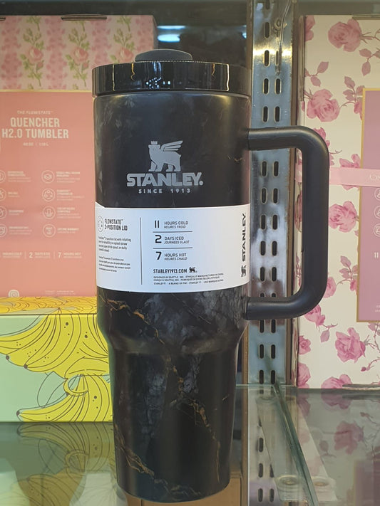 Stanley black tumbler with handle and lid displayed on a glass shelf with pink and yellow background