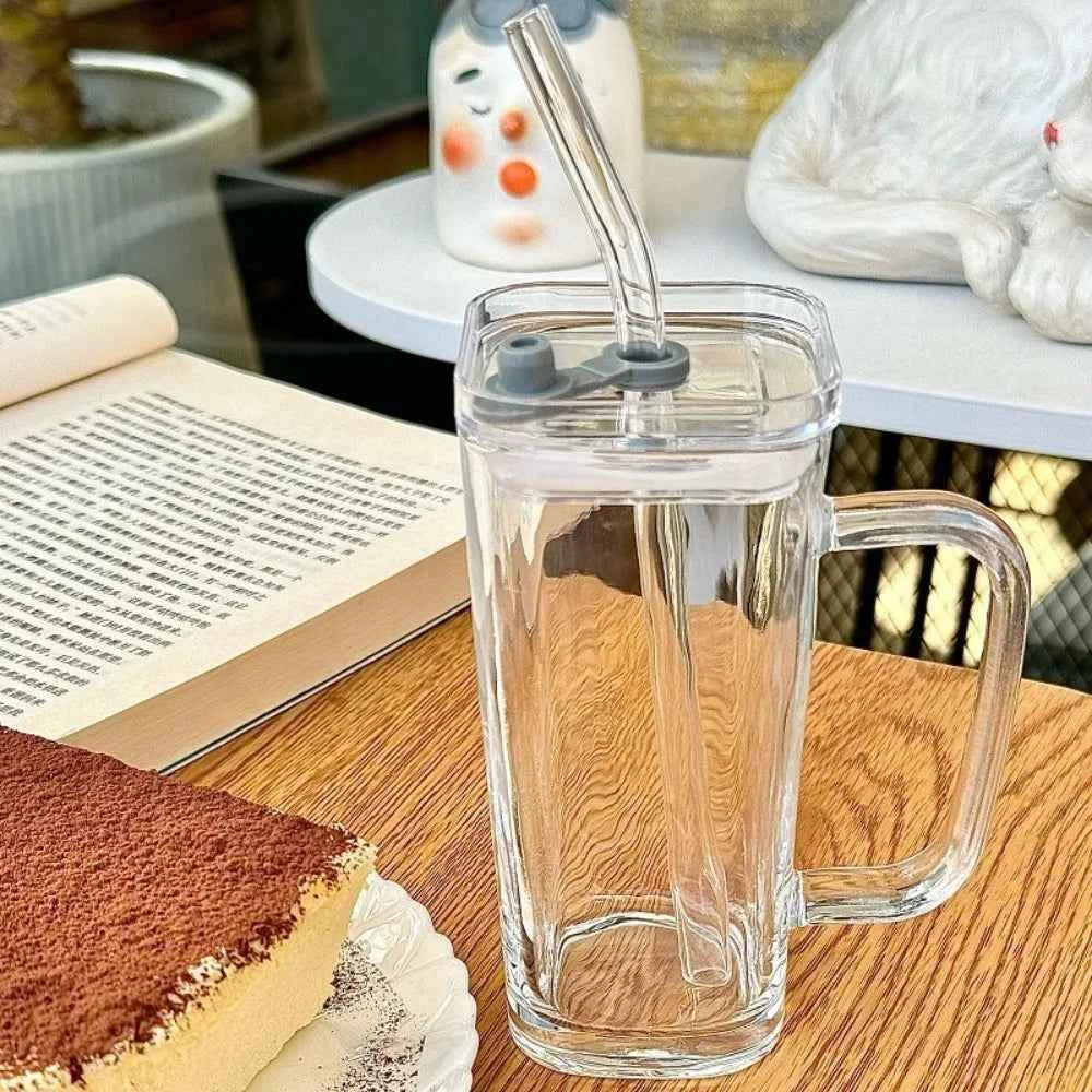 Stylish square glass mug with a straw on a wooden table beside a book and dessert