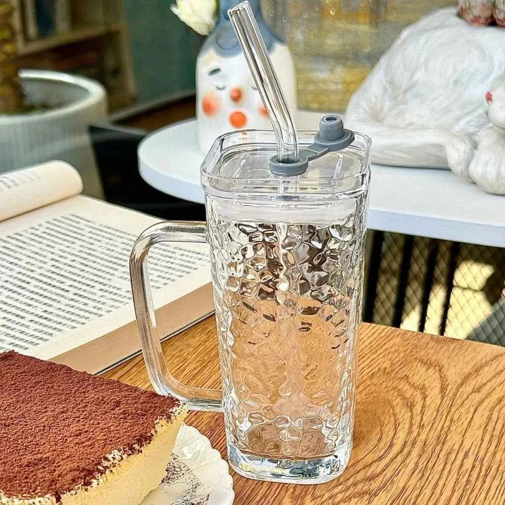 Stylish square glass mug with a handle and straw on a wooden table next to a dessert