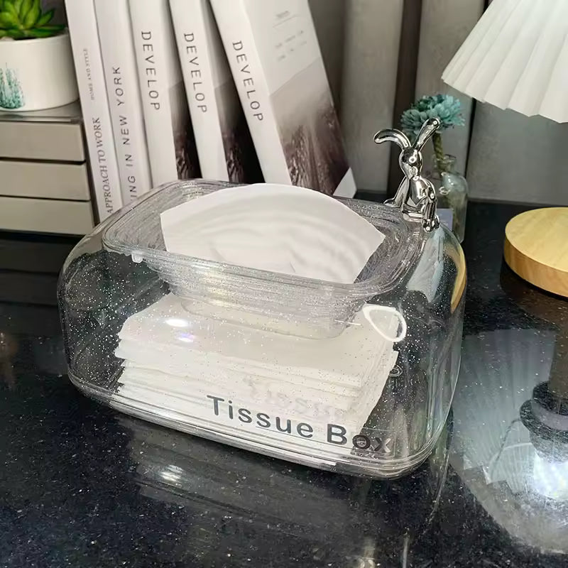 Clear sunken tissue box organizer with silver rabbit handle on black countertop by books and decor