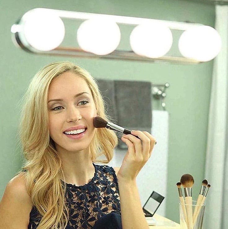 Woman applying makeup with brushes under a 4 bulb makeup mirror light mounted on the wall