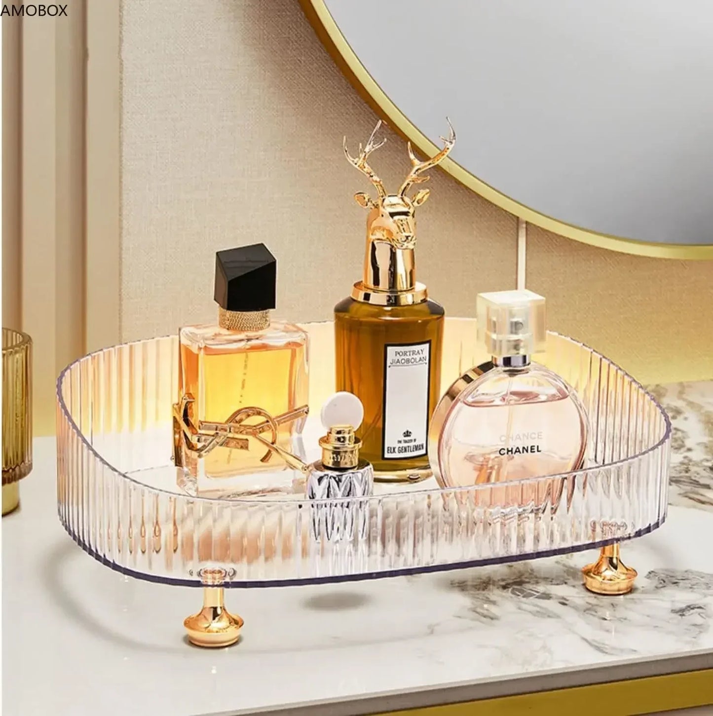 Clear transparent dresser storage box with gold feet holding assorted perfume bottles on marble countertop