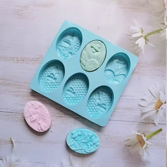 Clarify.pk 6 cavity oval soap mold in blue silicone with floral design and pastel soap bars on wooden surface
