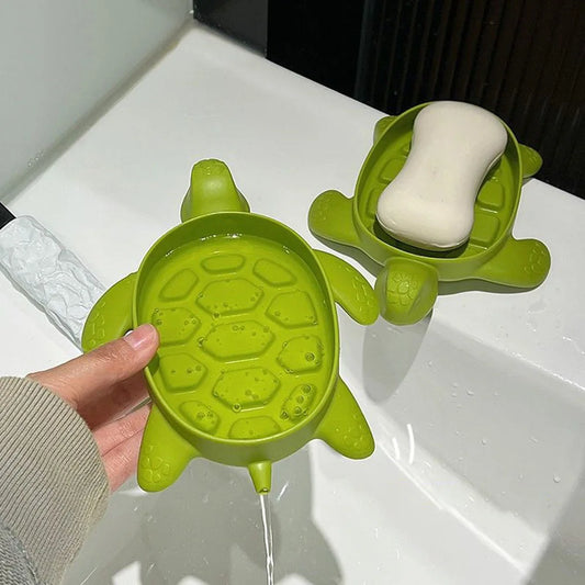 Green cartoon turtle soap holder with drainage feature holding white soap bar on bathroom sink