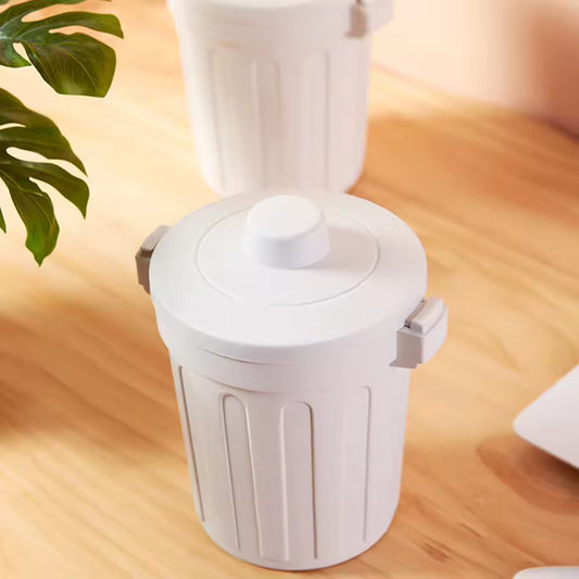 White desktop mini trash can with lid on wooden desk near green plant and laptop
