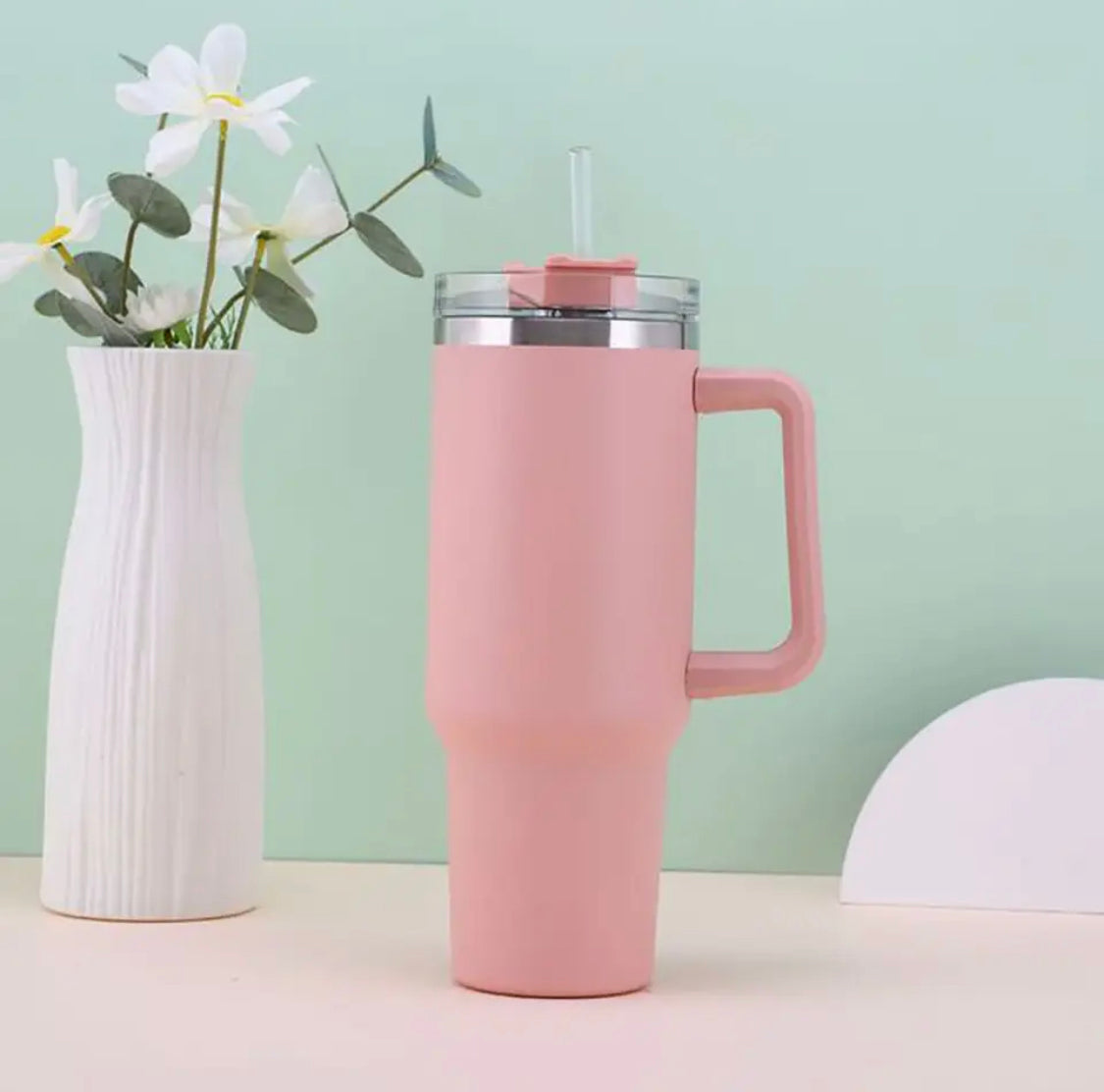 Pink double wall vacuum tumbler with handle and clear lid featuring straw beside white vase with flowers