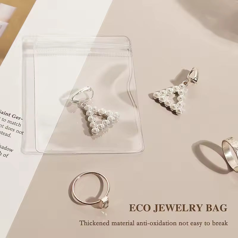 Clear eco jewelry bag with pearl triangle earrings and a silver ring on a beige surface