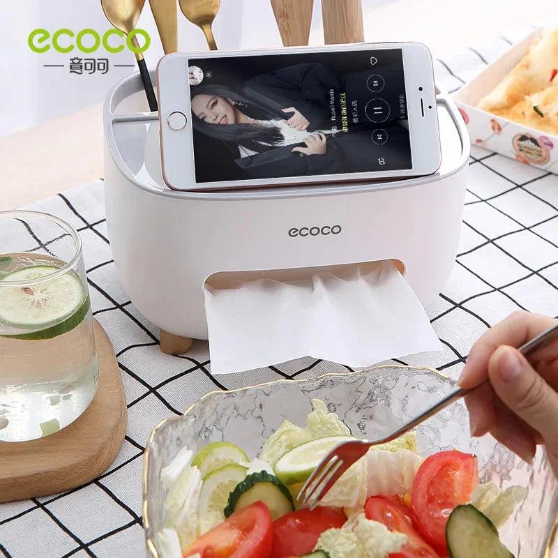 White Ecoco tissue box holder with phone stand showing a smartphone on top, placed on a dining table