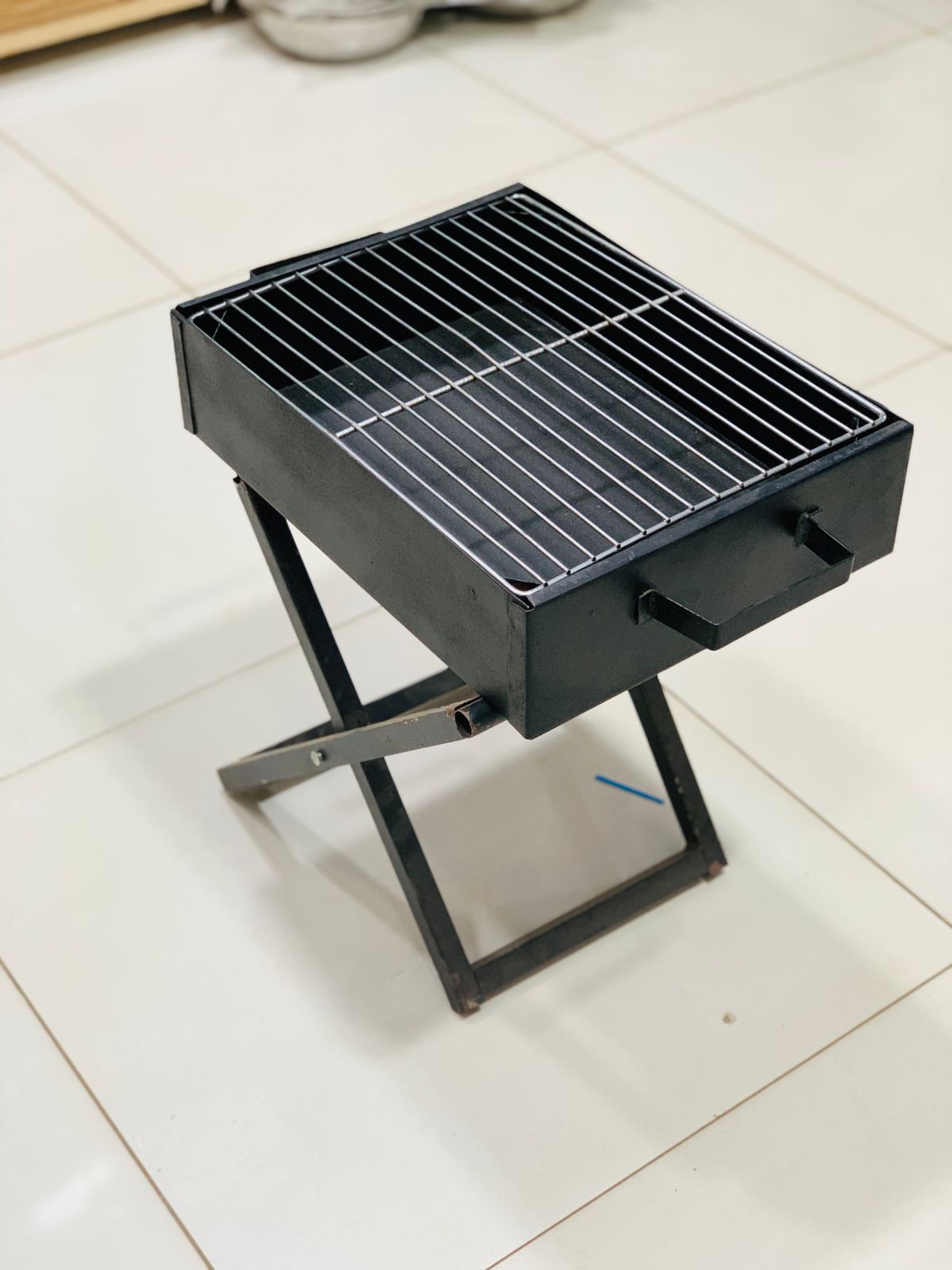Compact black bbq grill for home use with foldable legs and metal grilling surface on white tiled floor