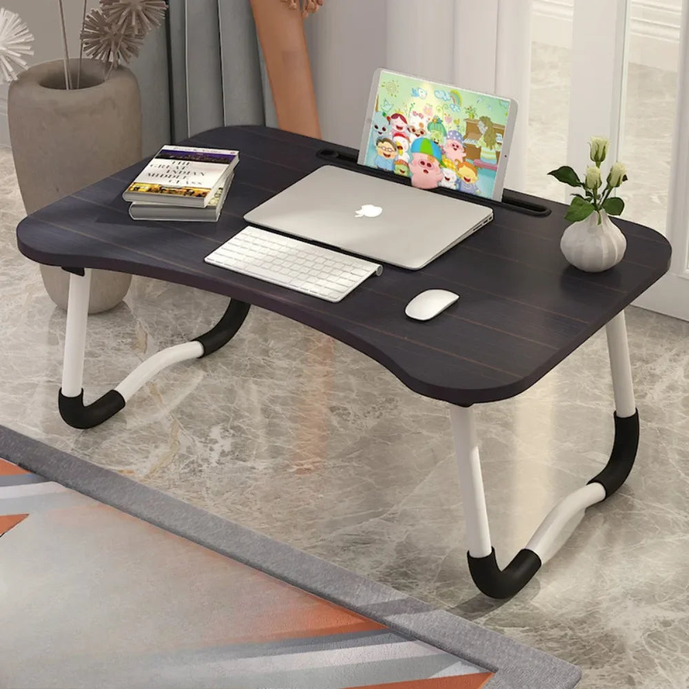 Dark wood foldable laptop desk with white curved legs holding a laptop, keyboard, mouse, books, and a plant