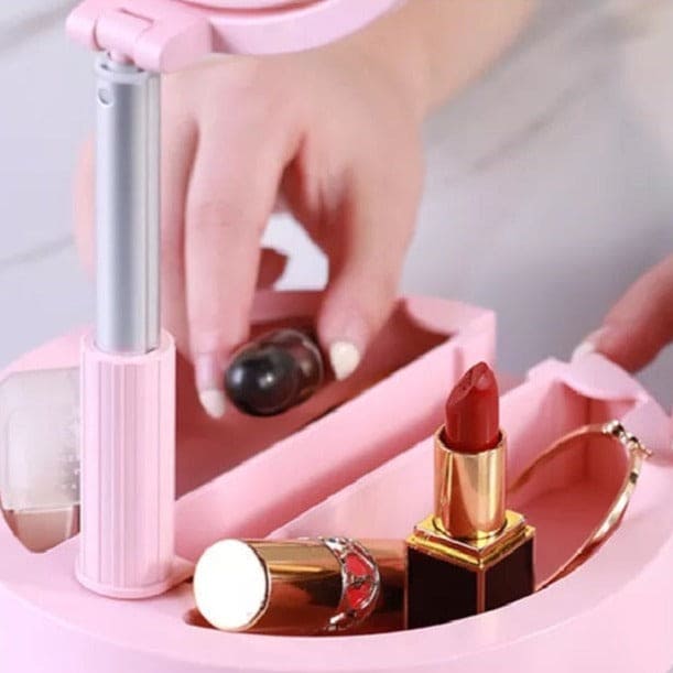 Pink foldable selfie stick with ring light placed next to cosmetics and hand adjusting lipstick
