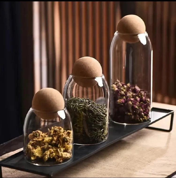 Set of three glass storage jars with cork lids containing dried herbs and flowers on a black tray