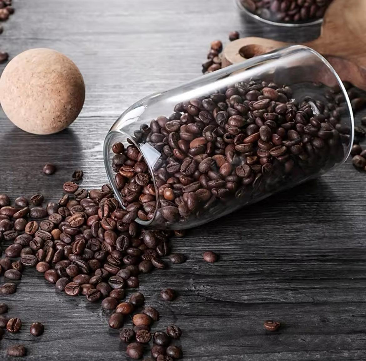 Clear glass storage jars with cork lids spilling coffee beans on a dark wooden surface