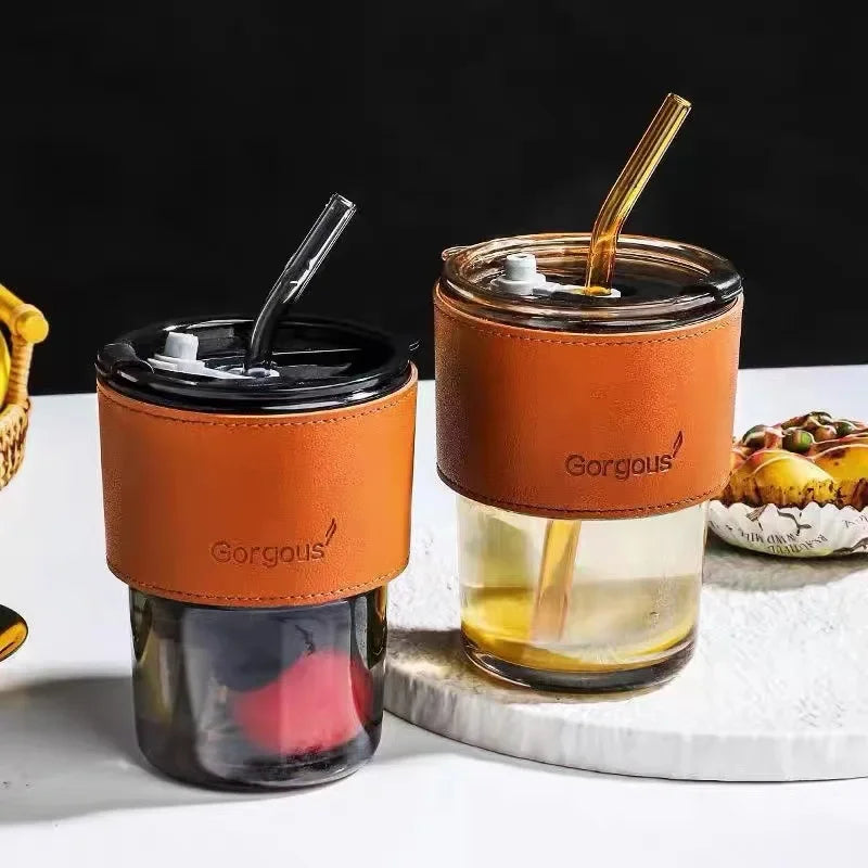 Two bamboo node glass straw cups with brown leather sleeves and matching lids on white surface