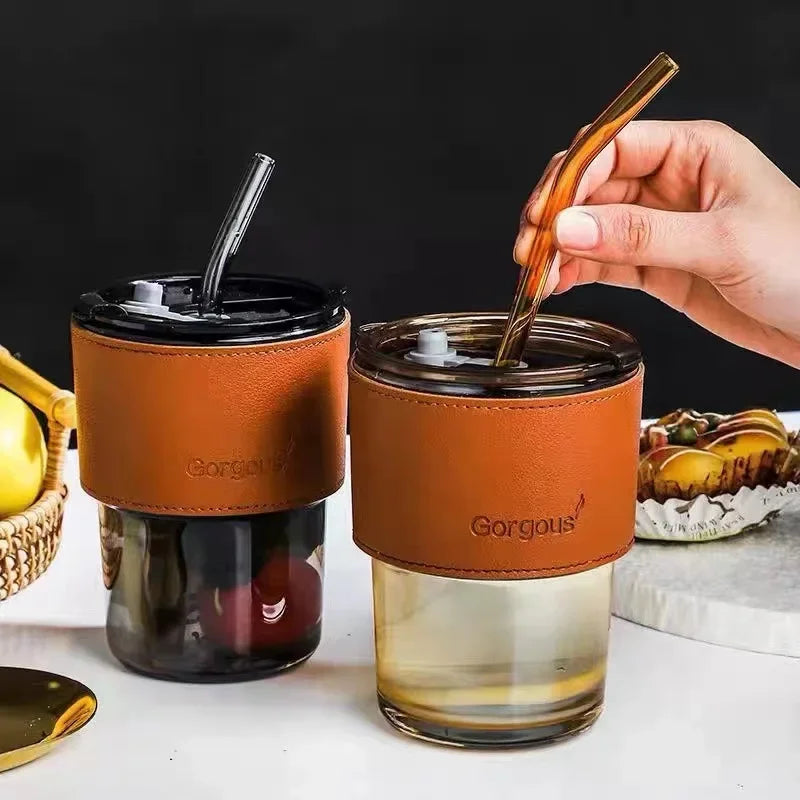 Bamboo node glass straw cup with brown leather sleeve and lids, one held by hand on white surface