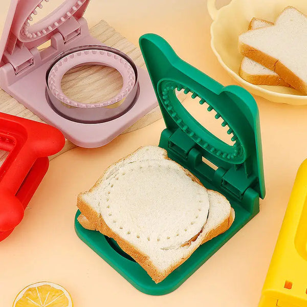 Green sandwich cutter and sealer pressing crustless white bread sandwich with pink red yellow cutters nearby
