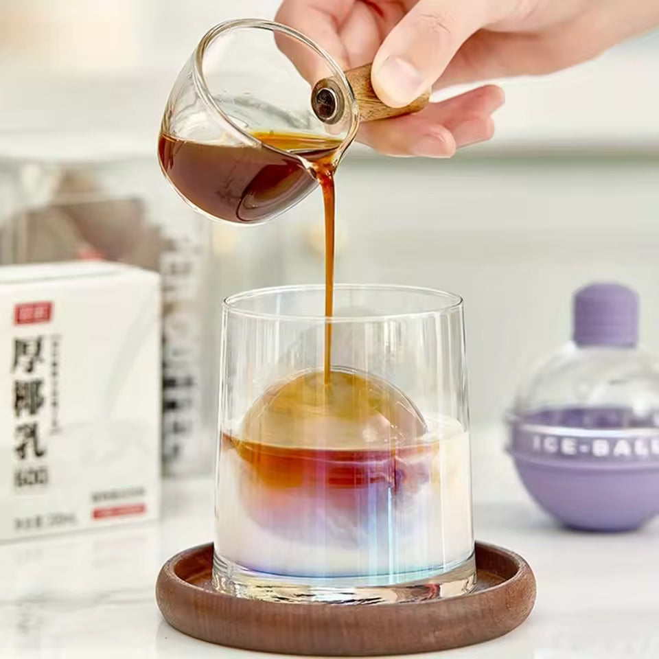 Glass of iced drink with spherical ice mold and coffee being poured from a small glass pitcher
