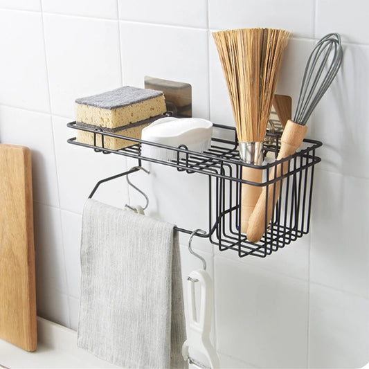 Black metal sink shelf organizer mounted on white tile wall holding sponge soap whisk broom and hanging towel