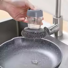 Hand holding a kitchen soap dispensing brush with steel scrubber cleaning a frying pan under faucet
