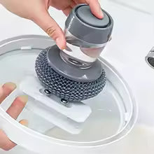 Gray kitchen soap dispensing brush with ergonomic handle being used to clean a glass lid