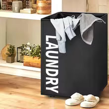 Black laundry storage basket by Clarify.pk with white text and clothes inside placed on wooden floor