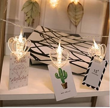 Clarify.pk LED photo clip string lights with crown-shaped clips holding decorative cards on a white shelf