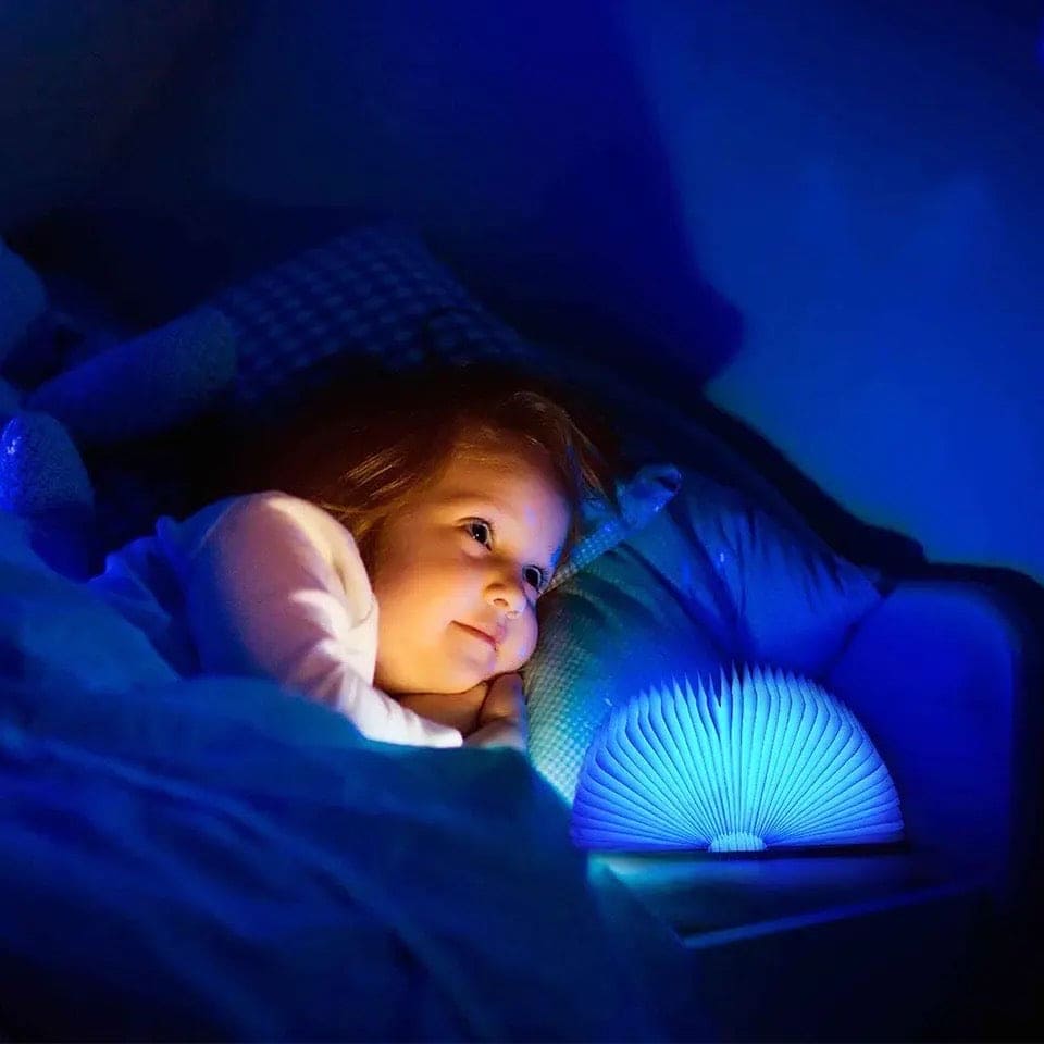 Child lying in bed next to a glowing blue LED wooden book lamp providing soft night light