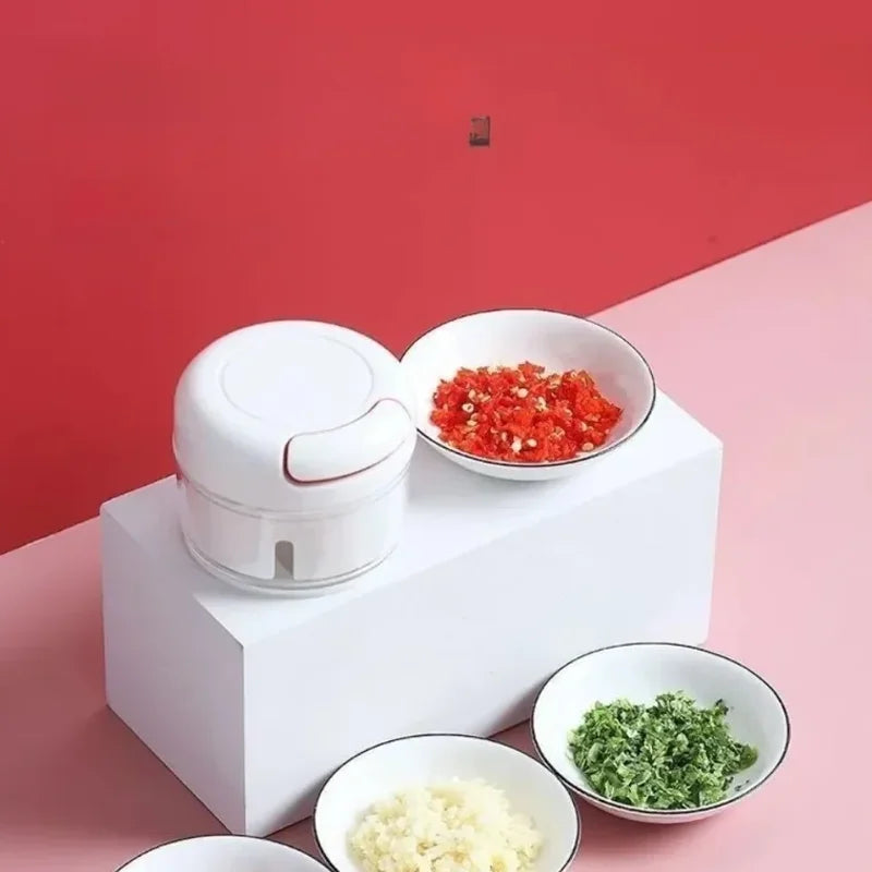 White manual garlic chopper placed on white block with chopped garlic, red peppers, and green herbs in bowls