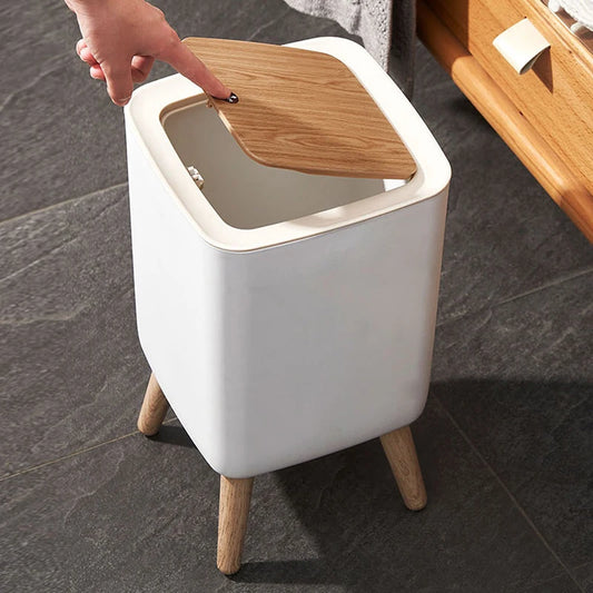 Modern square trash bin with wooden lid and legs on dark tile floor in home setting