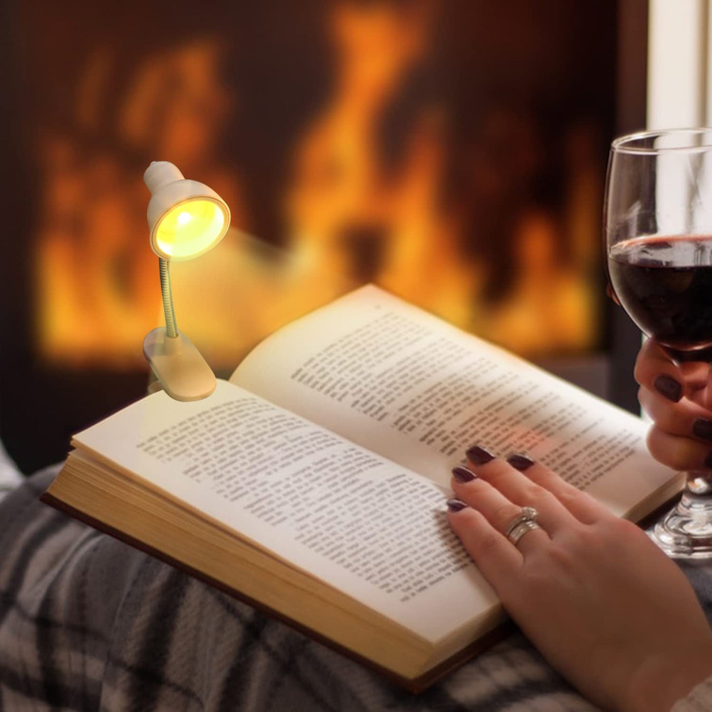 Portable book reading light clipped to a book illuminating pages by a cozy fireplace with a person holding a glass of wine