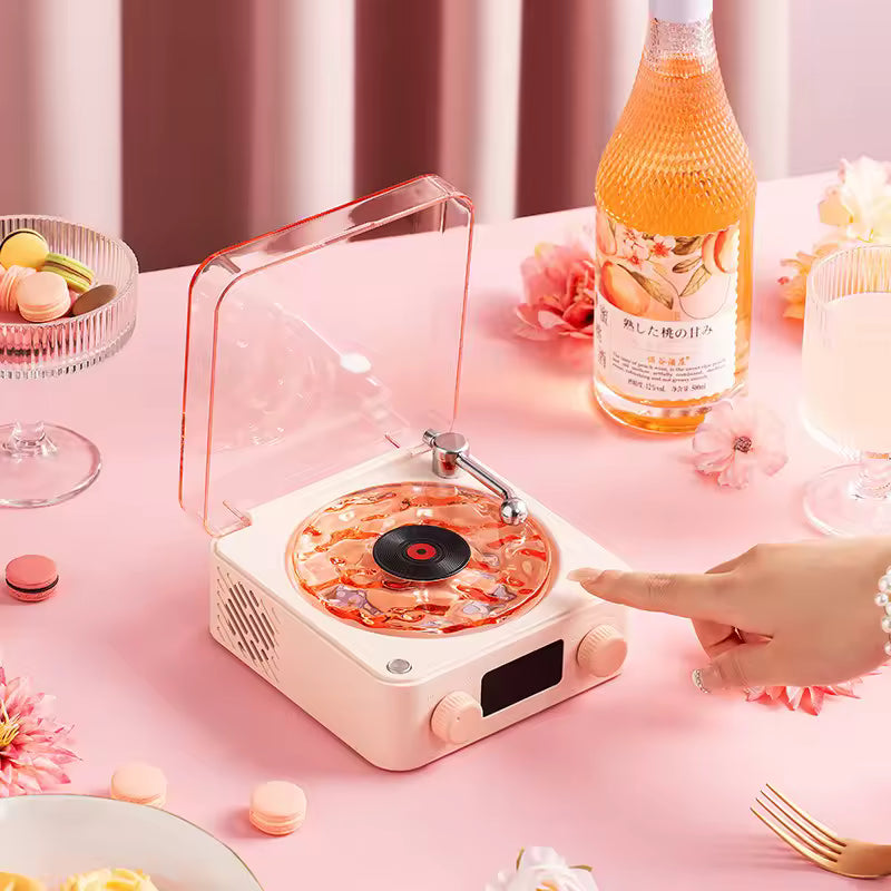 Retro turntable Bluetooth speaker in white with transparent cover on a pink table surrounded by desserts and drinks