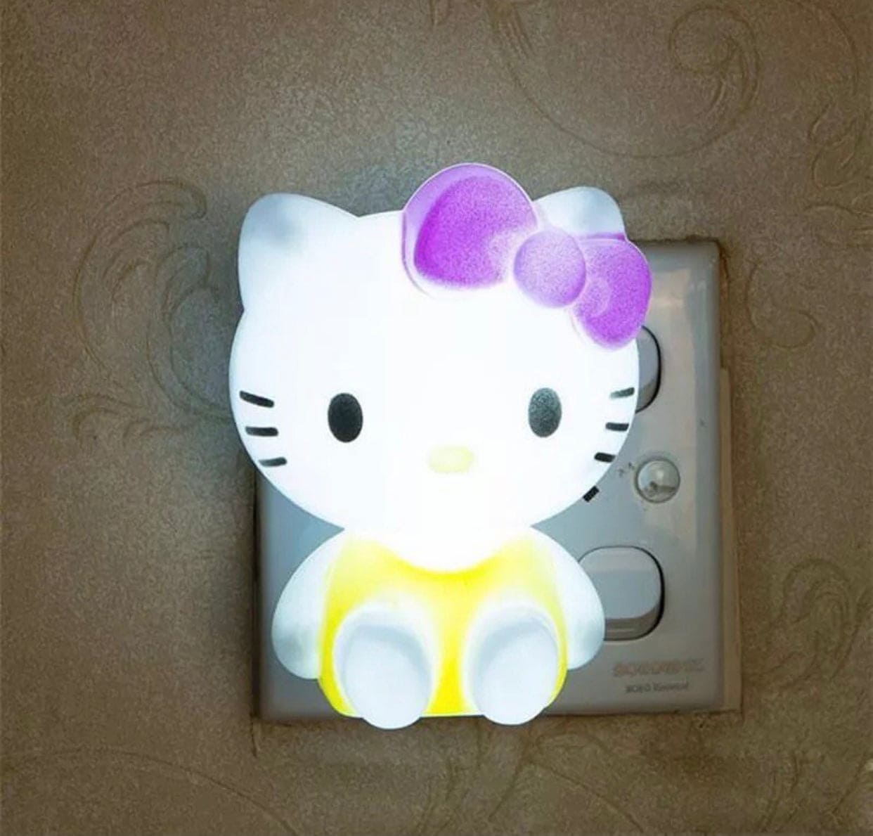 Hello Kitty night light plugged into a wall outlet glowing with purple bow and yellow outfit