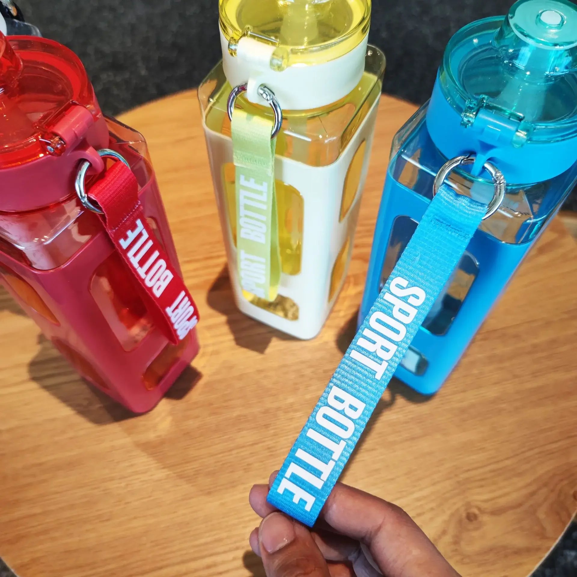 Three colorful 900ml leak proof water bottles in red, yellow, and blue with carrying straps on a wooden surface