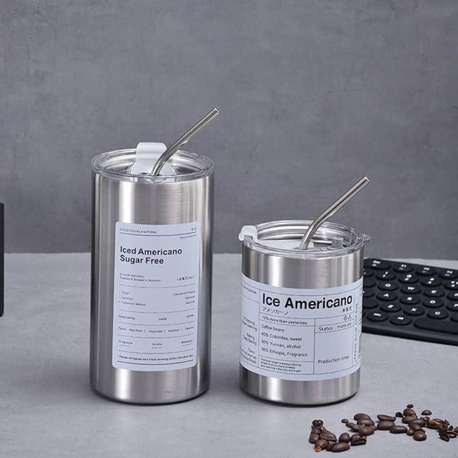 Two stainless steel coffee tumblers with lids and metal straws on a gray surface with coffee beans nearby