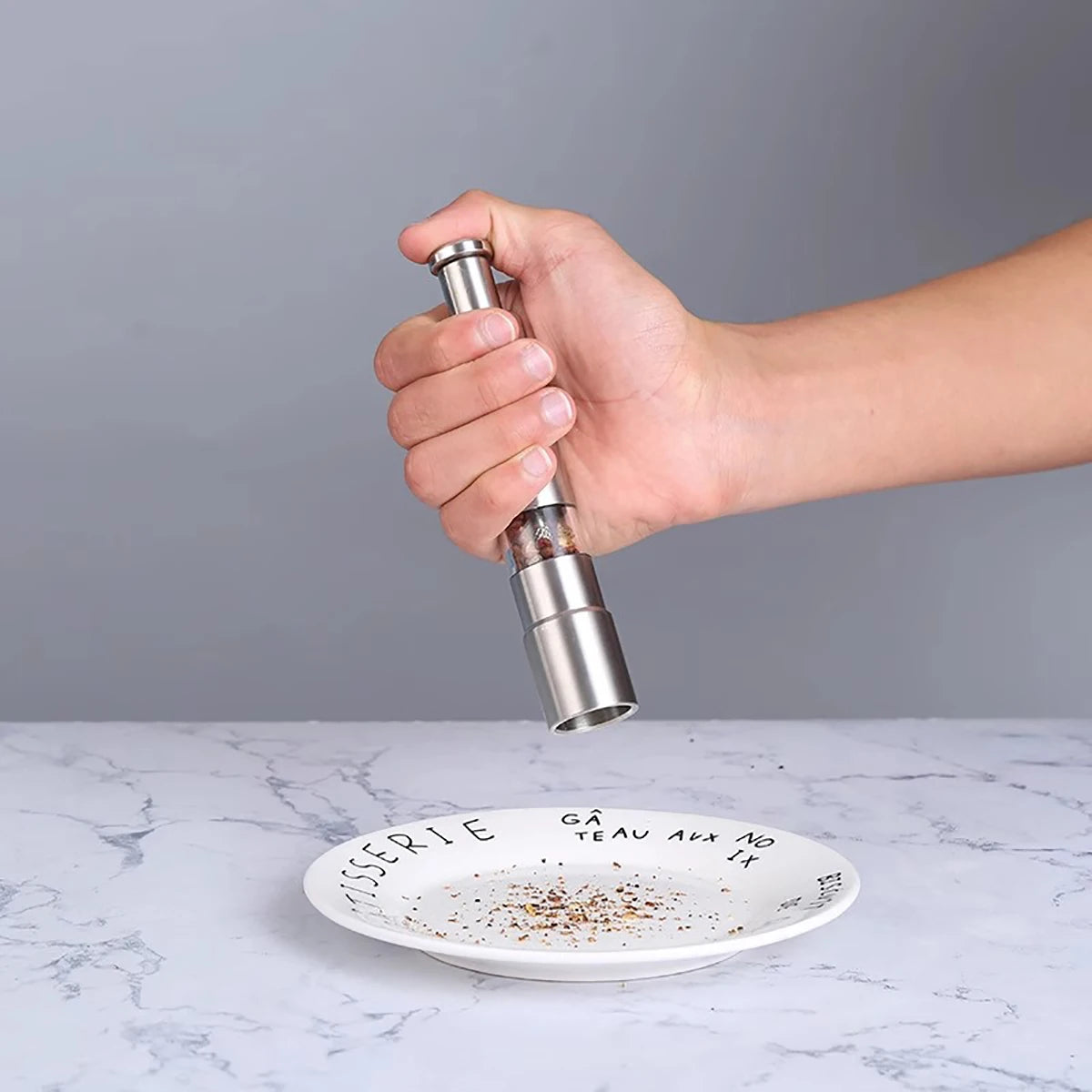 Hand holding a stainless steel pepper grinder seasoning a white plate on marble countertop