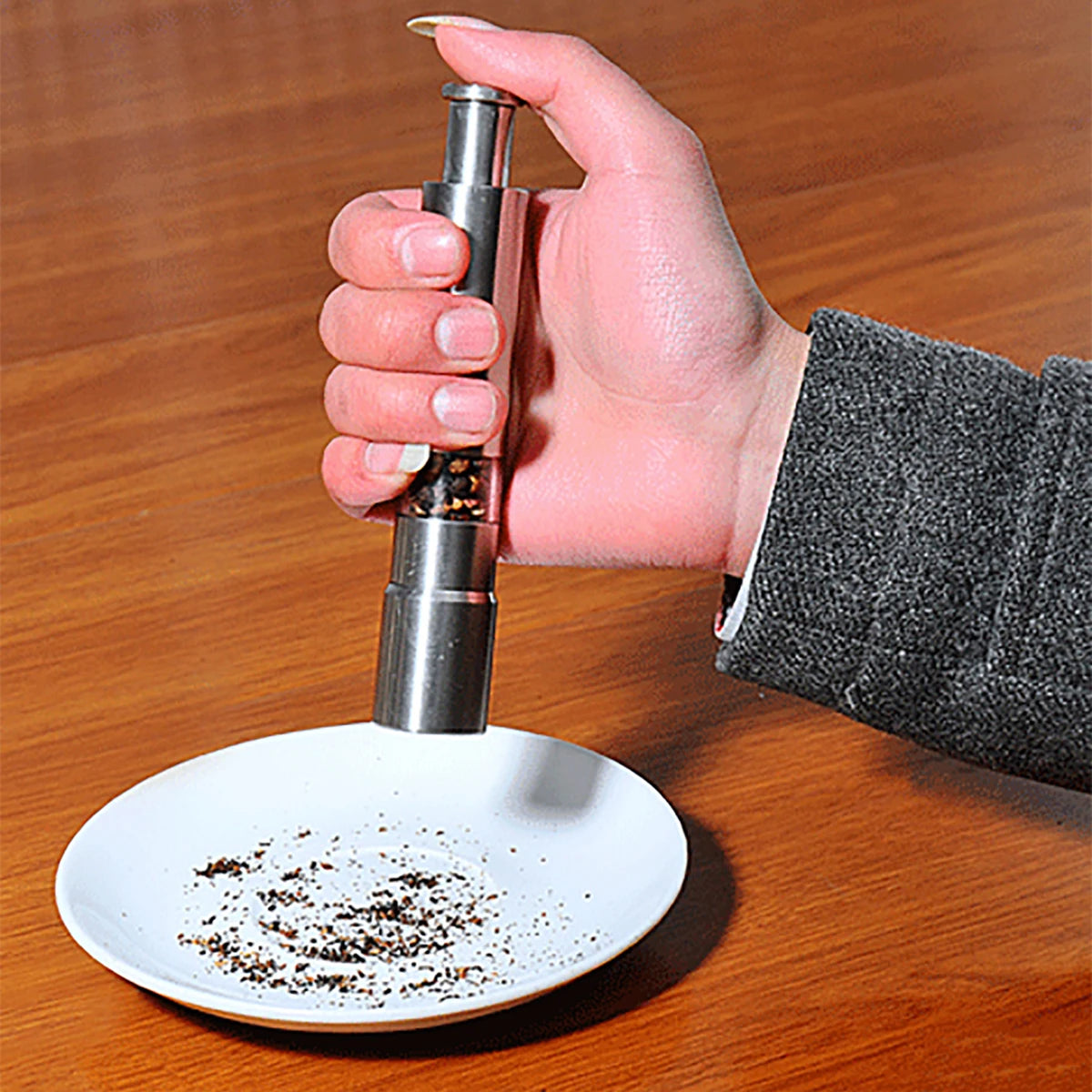 Hand using stainless steel pepper grinder to grind pepper over a white plate on wooden surface