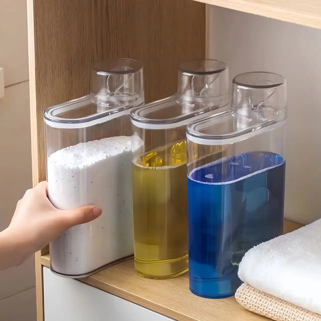 Hand using a transparent laundry detergent dispenser set with liquid and powder detergent containers on a wooden shelf